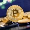 A close-up of a stack of gold-colored Bitcoin coins, with one coin standing upright and a blurred digital stock chart in the background.