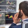 A man in a suit talks on the phone while looking at multiple computer screens displaying colorful stock market charts and graphs.
