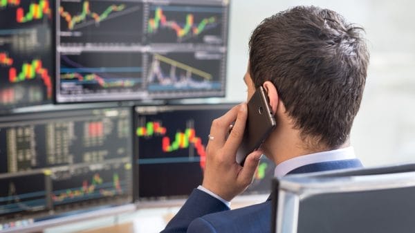 A man in a suit talks on the phone while looking at multiple computer screens displaying colorful stock market charts and graphs.
