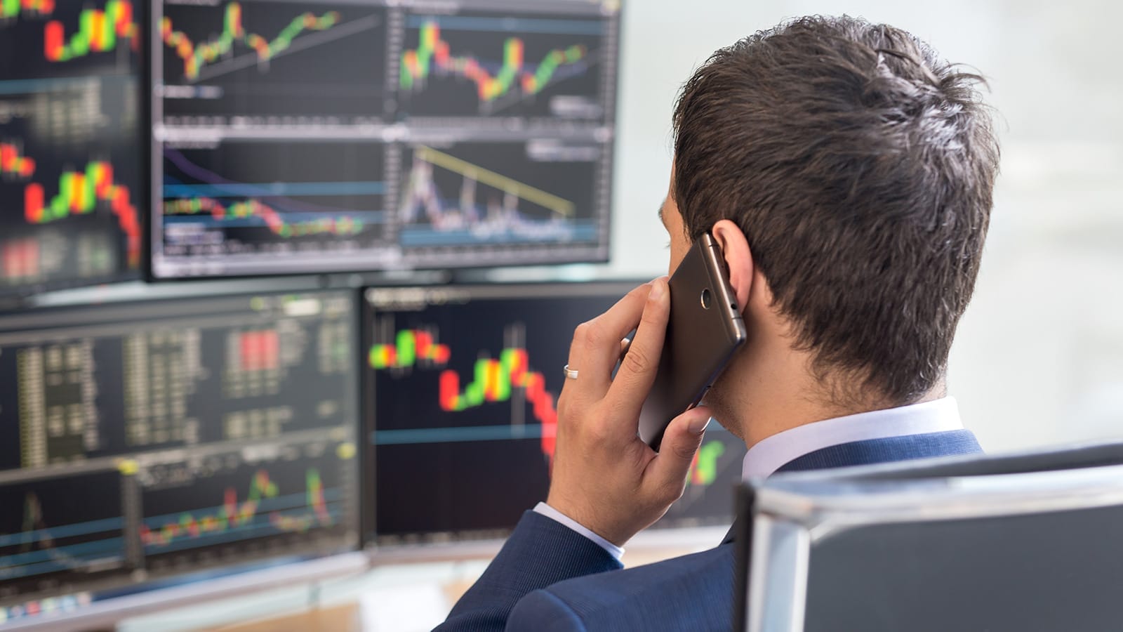 A man in a suit talks on the phone while looking at multiple computer screens displaying colorful stock market charts and graphs.