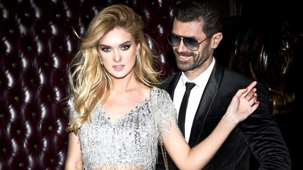 A woman in a sparkly silver dress stands confidently beside a man in a black suit and sunglasses, both posing stylishly against a dark, tufted leather background.