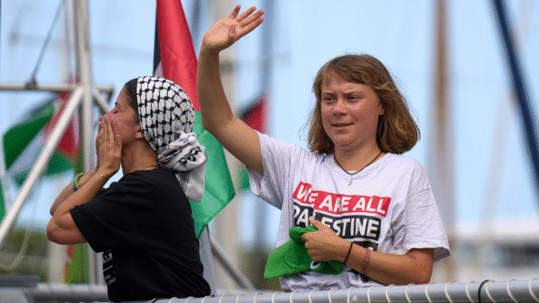 Two women on a boat; one waves while the other blows a kiss. Both wear "We are all Palestine" shirts and headscarves, with Palestinian flags visible in the background.