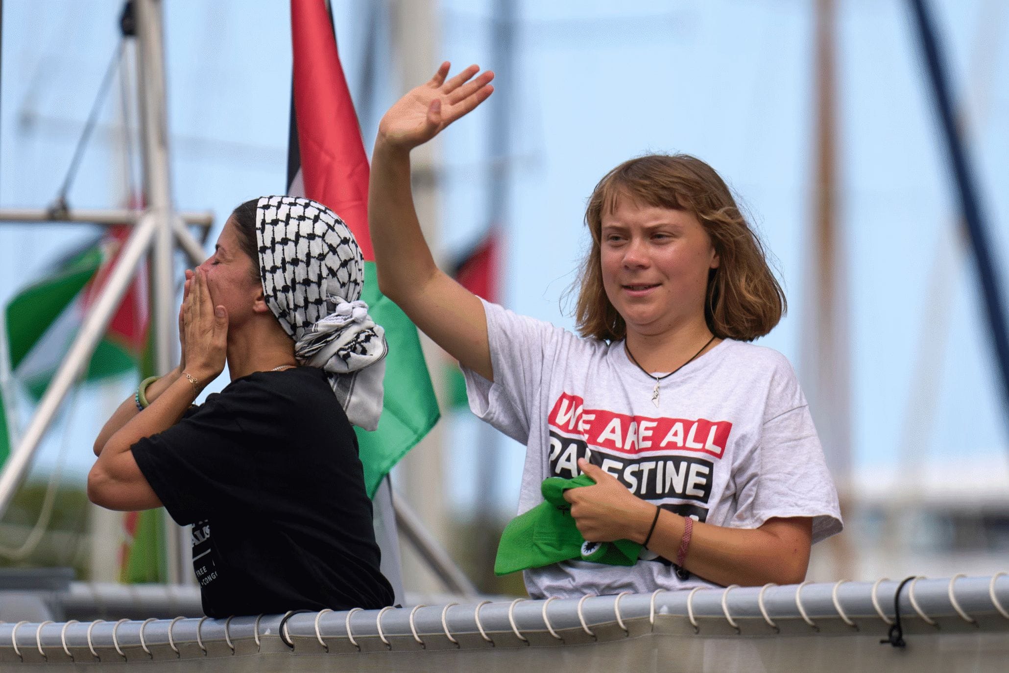 Two women on a boat; one waves while the other blows a kiss. Both wear "We are all Palestine" shirts and headscarves, with Palestinian flags visible in the background.