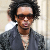A man with curly hair, sunglasses, and layered necklaces wears a patterned black jacket with a satin lapel, standing outdoors.