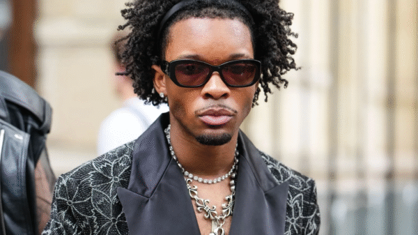 A man with curly hair, sunglasses, and layered necklaces wears a patterned black jacket with a satin lapel, standing outdoors.