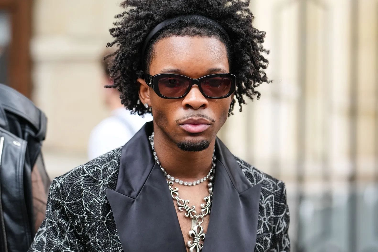 A man with curly hair, sunglasses, and layered necklaces wears a patterned black jacket with a satin lapel, standing outdoors.