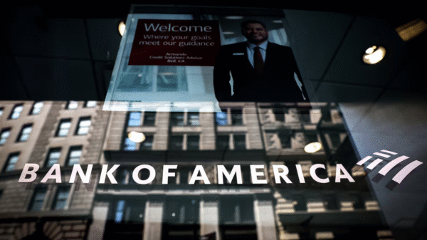 Bank of America sign on a glass window, reflecting city buildings, with a poster of a smiling advisor inside.
