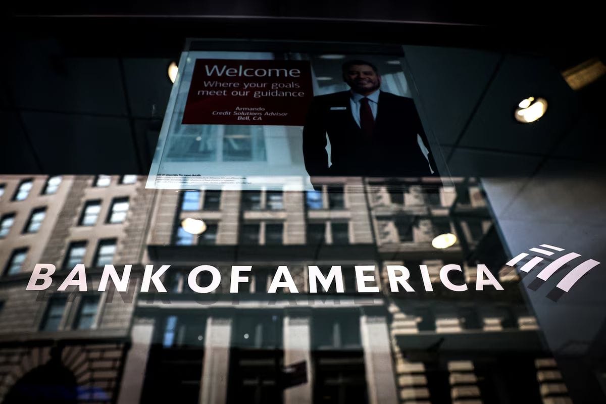 Bank of America sign on a glass window, reflecting city buildings, with a poster of a smiling advisor inside.