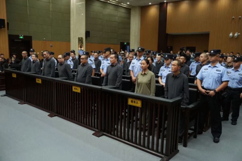 A group of people stands in a courtroom behind a wooden barrier, facing forward, with uniformed police officers standing behind them.