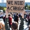 A person holds a cardboard sign reading "WE NEED A CHANGE" above their head in a crowd at an outdoor protest or rally.