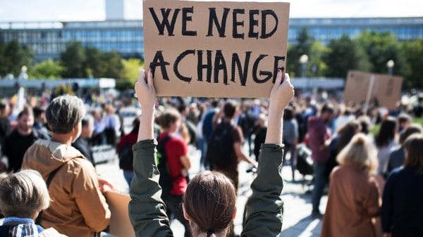 A person holds a cardboard sign reading "WE NEED A CHANGE" above their head in a crowd at an outdoor protest or rally.