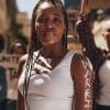 A woman stands confidently at a protest with “WARRIOR” painted on her arm, while others hold signs in the background.