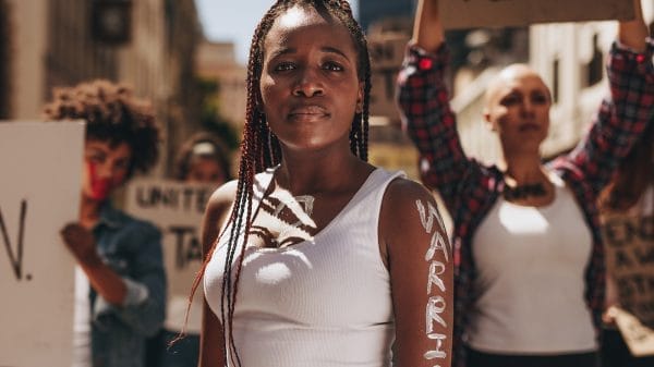 A woman stands confidently at a protest with “WARRIOR” painted on her arm, while others hold signs in the background.