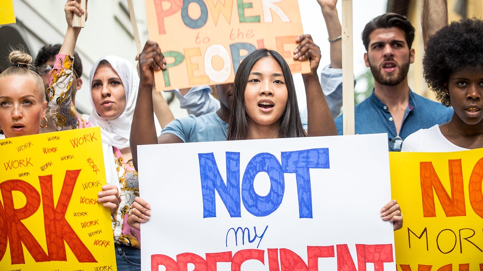 A diverse group of protesters hold signs, including one reading "NOT my PRESIDENT," while expressing their opinions at a rally.