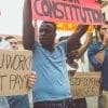 A diverse group of people at a protest hold signs with messages about fair wages and rights, including “WOULD U WORK WITHOUT PAY?” and “POWER TO THE PEOPLE.”.