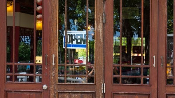 A glass door with a hanging “OPEN” sign and a yellow “PULL” label, showing reflections of trees and a glimpse of the interior.