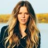 A woman with long wavy blonde hair stands outdoors by a lake, wearing a dark jacket, with trees and water blurred in the background.