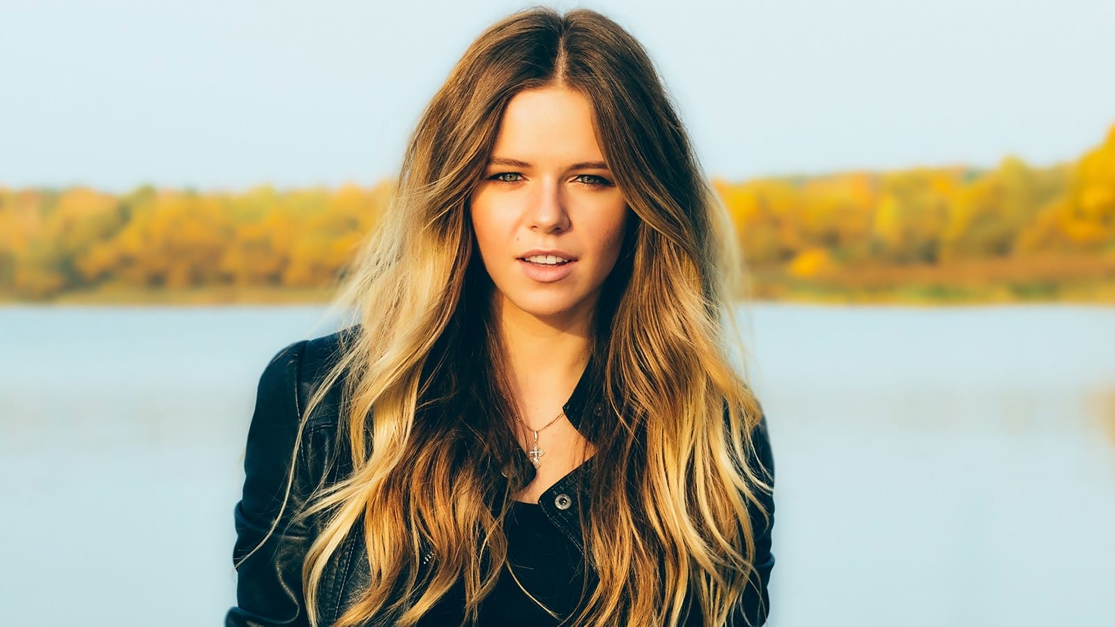 A woman with long wavy blonde hair stands outdoors by a lake, wearing a dark jacket, with trees and water blurred in the background.