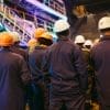 A group of workers in uniforms and hard hats stand together in an industrial facility, facing away from the camera.