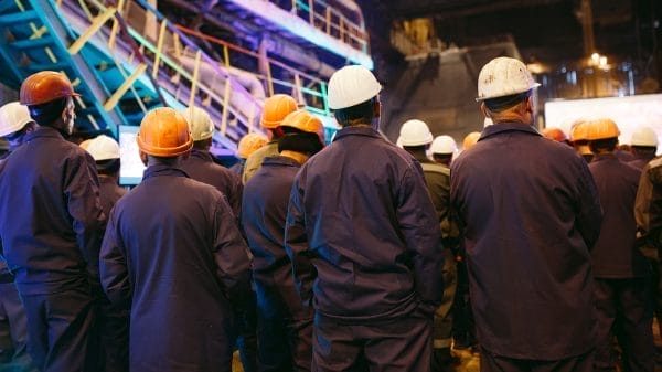 A group of workers in uniforms and hard hats stand together in an industrial facility, facing away from the camera.