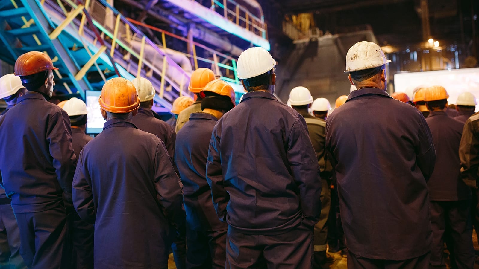 A group of workers in uniforms and hard hats stand together in an industrial facility, facing away from the camera.