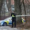 A police officer stands by police tape near a wooded area, with a police car parked nearby on a cloudy day.
