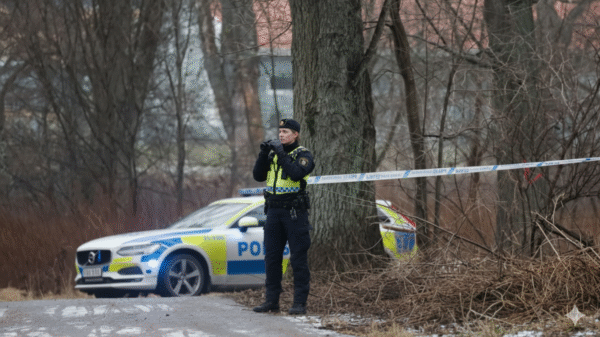 A police officer stands by police tape near a wooded area, with a police car parked nearby on a cloudy day.