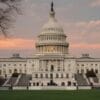 The United States Capitol building at sunset, with a pink-orange sky and trees framing the foreground.