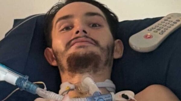 A young man with a beard lies in a hospital bed, using a ventilator tube. A remote control rests on the pillow beside him.