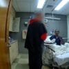 A person in a hospital bed talks to another standing person in a small hospital room with medical equipment and cabinets.