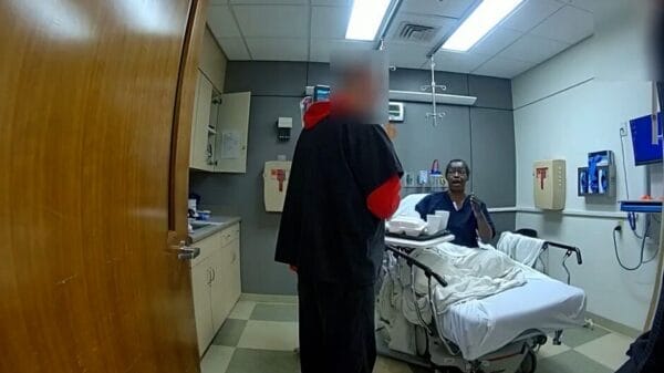 A person in a hospital bed talks to another standing person in a small hospital room with medical equipment and cabinets.