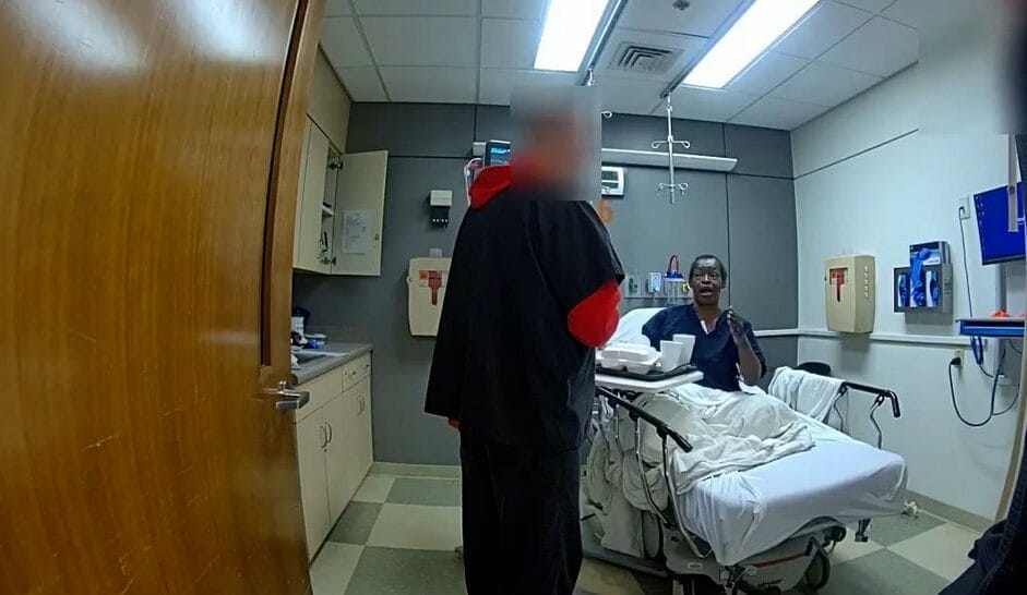 A person in a hospital bed talks to another standing person in a small hospital room with medical equipment and cabinets.