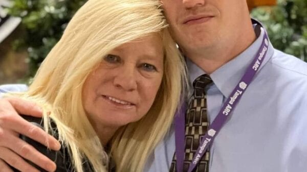 A woman and a man pose together, hugging and smiling, with greenery and an "ARMY" sign visible in the background.