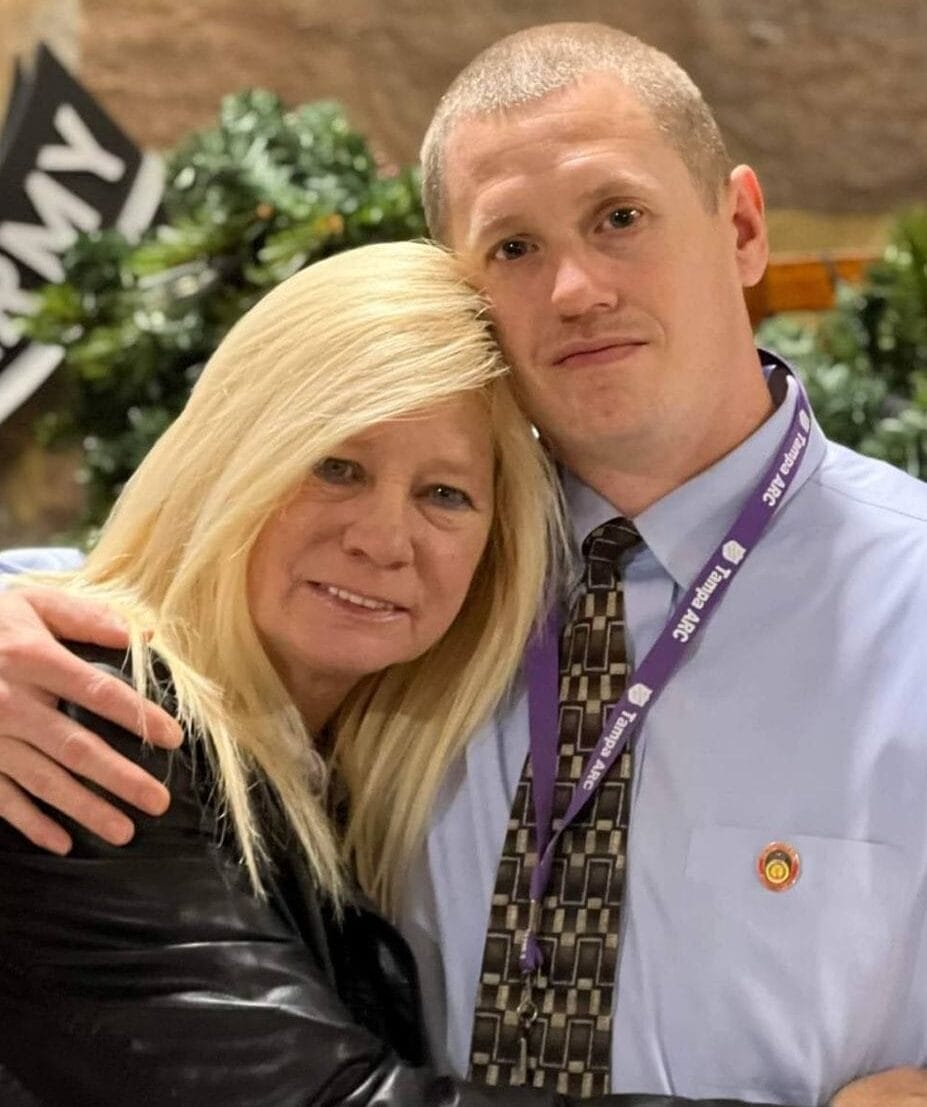 A woman and a man pose together, hugging and smiling, with greenery and an "ARMY" sign visible in the background.