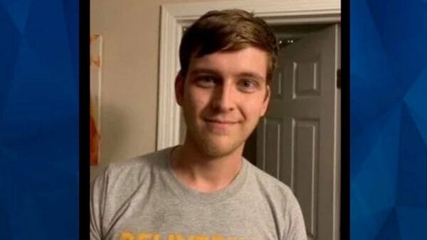 A young man with short brown hair stands indoors, smiling at the camera. He is wearing a gray T-shirt with yellow writing. A white door is open in the background.