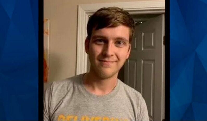 A young man with short brown hair stands indoors, smiling at the camera. He is wearing a gray T-shirt with yellow writing. A white door is open in the background.