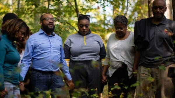 A group of five people stand close together outdoors in a wooded area, holding hands and appearing solemn or thoughtful.