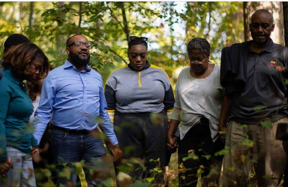 A group of five people stand close together outdoors in a wooded area, holding hands and appearing solemn or thoughtful.