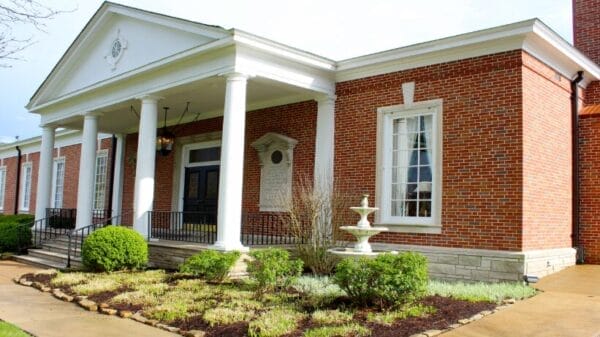 A red-brick building with white columns, large windows, a black double door, trimmed bushes, a small white fountain, and a sidewalk in front.