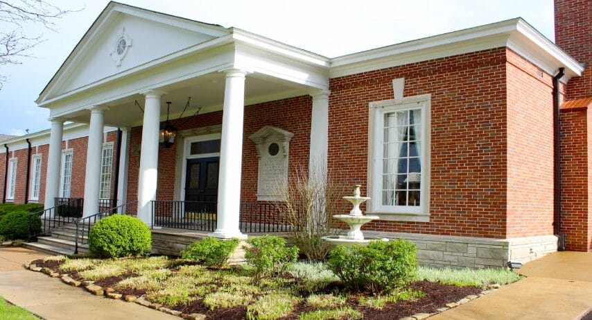 A red-brick building with white columns, large windows, a black double door, trimmed bushes, a small white fountain, and a sidewalk in front.