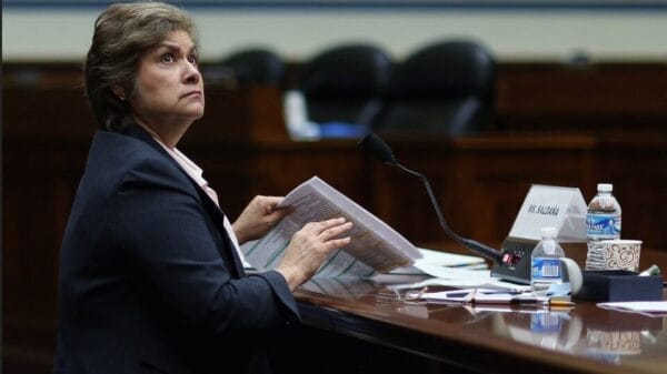 A woman in a suit sits at a desk with papers and a microphone, looking up. A nameplate, water bottles, and coffee cups are on the desk in a formal meeting room.
