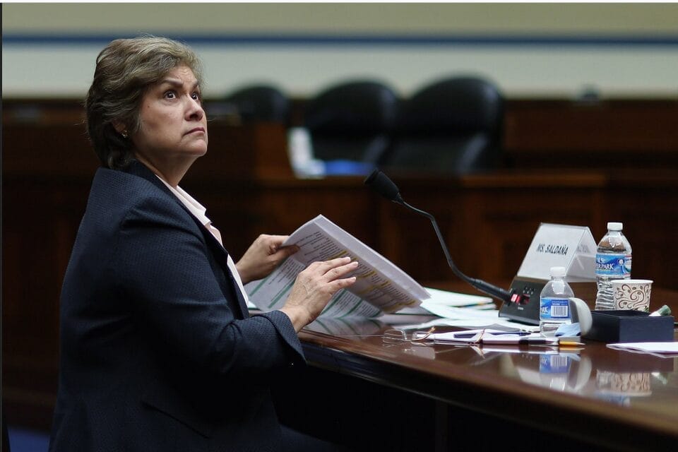 A woman in a suit sits at a desk with papers and a microphone, looking up. A nameplate, water bottles, and coffee cups are on the desk in a formal meeting room.