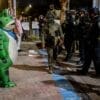 A person in a green frog costume faces a line of police officers in riot gear during a nighttime protest on a city street.