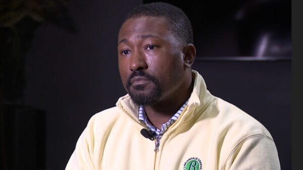 A man with a neatly trimmed beard wearing a light yellow zip-up jacket and plaid shirt sits indoors, appearing thoughtful.