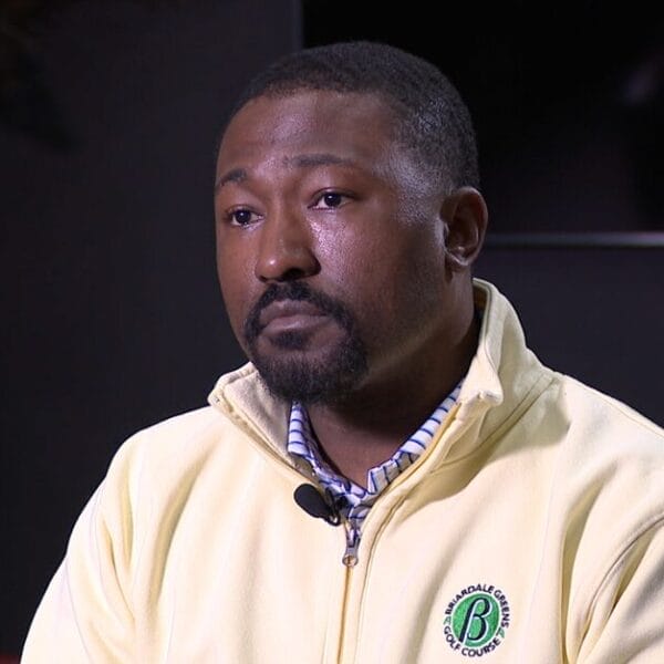 A man with a neatly trimmed beard wearing a light yellow zip-up jacket and plaid shirt sits indoors, appearing thoughtful.