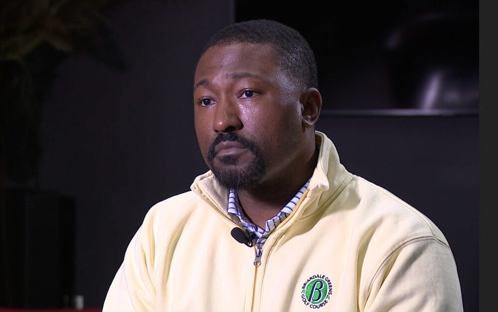 A man with a neatly trimmed beard wearing a light yellow zip-up jacket and plaid shirt sits indoors, appearing thoughtful.