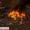 A white pickup truck engulfed in flames amid several burnt-out vehicles and debris, with thick black smoke rising. A "BREAKING NEWS" banner is at the bottom of the image.