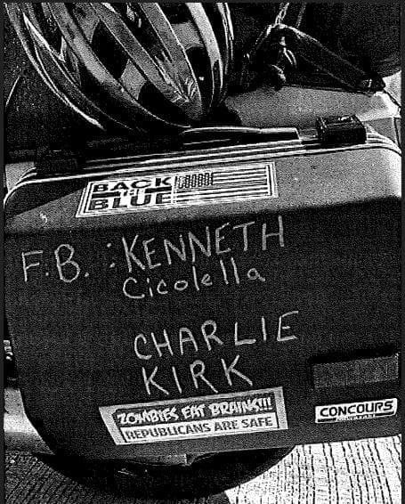 A motorcycle case with stickers and handwritten names: "F.B.: Kenneth Cicolella" and "Charlie Kirk." Stickers include "BACK THE BLUE" and "ZOMBIES EAT BRAINS!!! REPUBLICANS ARE SAFE.