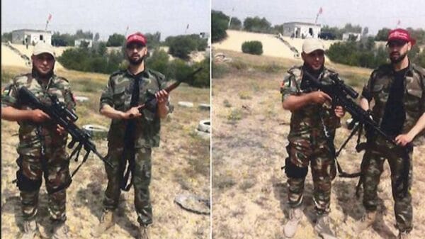 Two men wearing camouflage uniforms and caps stand outdoors on grassy terrain, each holding a rifle. Buildings and trees are visible in the background under a clear sky.