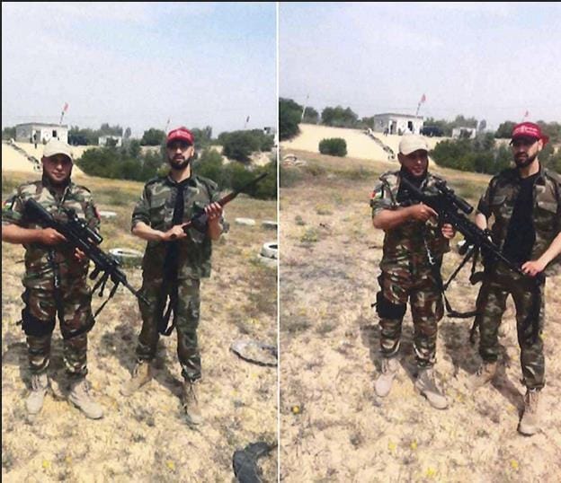 Two men wearing camouflage uniforms and caps stand outdoors on grassy terrain, each holding a rifle. Buildings and trees are visible in the background under a clear sky.